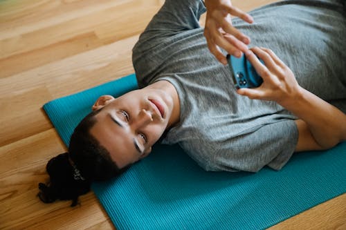 From above of young man in sportswear using mobile phone while lying on sports mat on floor at home