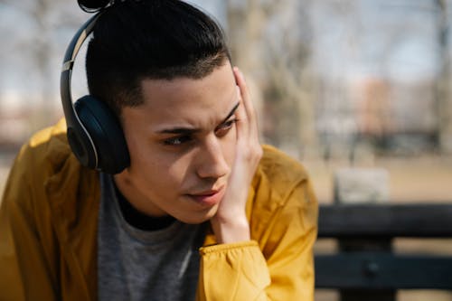 Free Thoughtful teen boy sitting on bench and listening to music in headphones while looking away on blurred background Stock Photo