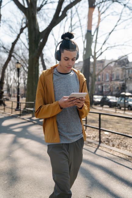 Male teenager in casual clothes browsing tablet while listening to music with headphones and walking in sunny autumn park with bare trees Male teenager in casual clothes browsing tablet while listening to music with headphones and walking in sunny autumn park with bare trees