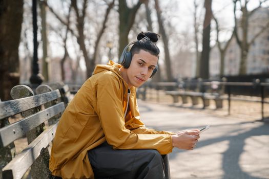 Side view of teen boy looking at camera and listening to music from smartphone while sitting on bench in autumn park with bare trees