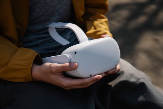 Crop anonymous person holding white VR headset in hands on blurred background in daylight