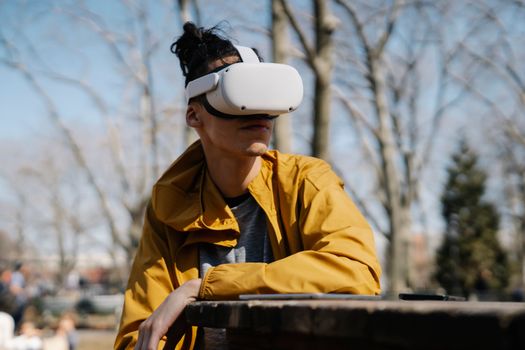Young male with long hair in bright yellow jacket sitting at wooden table while using VR goggles in park in autumn