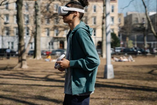 Side view of young male in casual outfit walking on street while using goggles of virtual reality and controllers in autumn