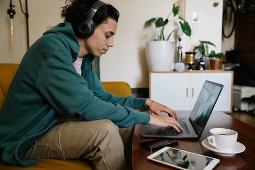 Side view of ethnic male distance employee in headset working on netbook at table with laptop and coffee in room