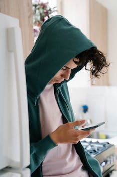 A young man wearing a hoodie focuses on his smartphone screen indoors, embodying modern digital lifestyle.