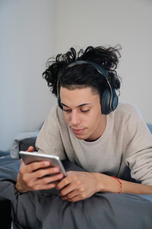 Interested young ethnic male listening to music from headset while lying on bed with cellphone in house
