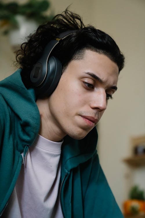 Concentrated young ethnic male in modern headset listening to music while looking down in house room