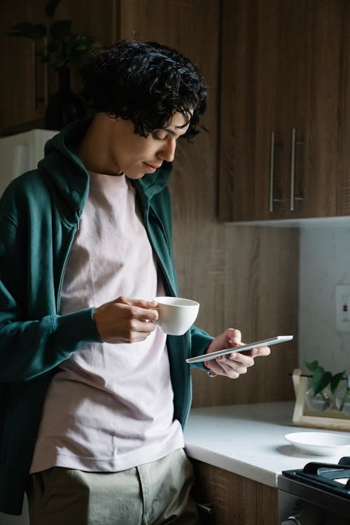 Attentive ethnic male with cup of hot drink text messaging on cellphone at home in daytime