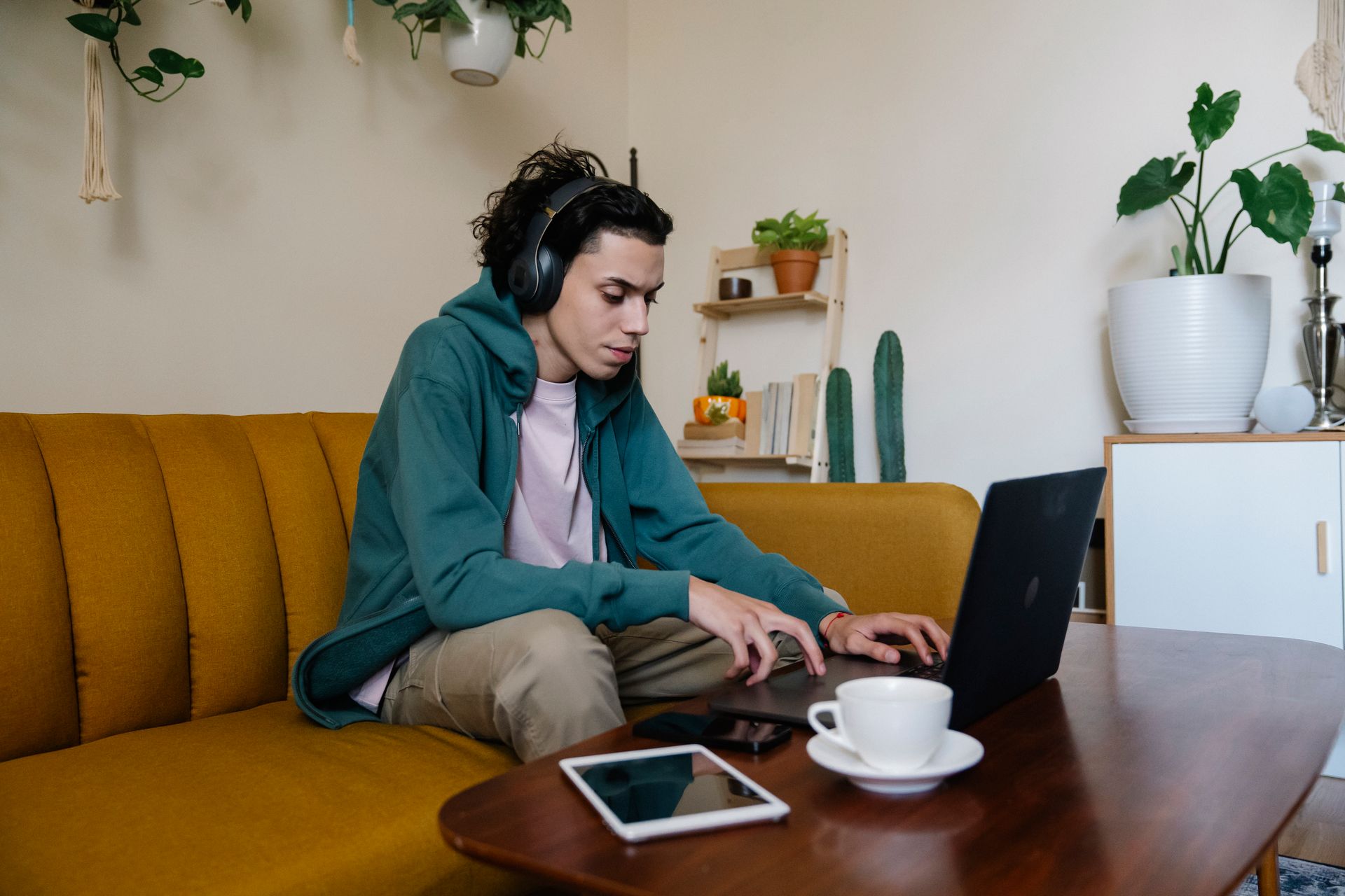 Attentive ethnic male remote worker in headset browsing internet on netbook while sitting at desk with tablet and hot drink in house
