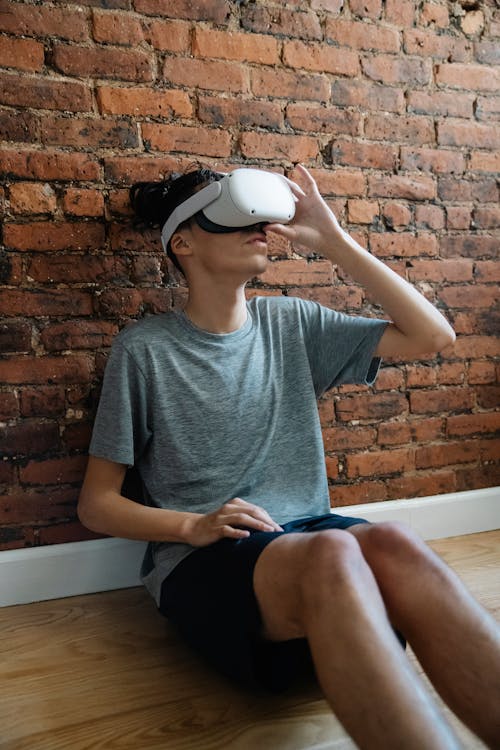 Male experiencing virtual reality in goggles while sitting on parquet against brick wall at home