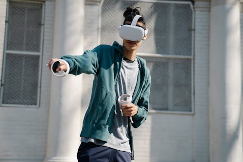 Man in virtual reality goggles with controllers playing video game against urban building in sunlight