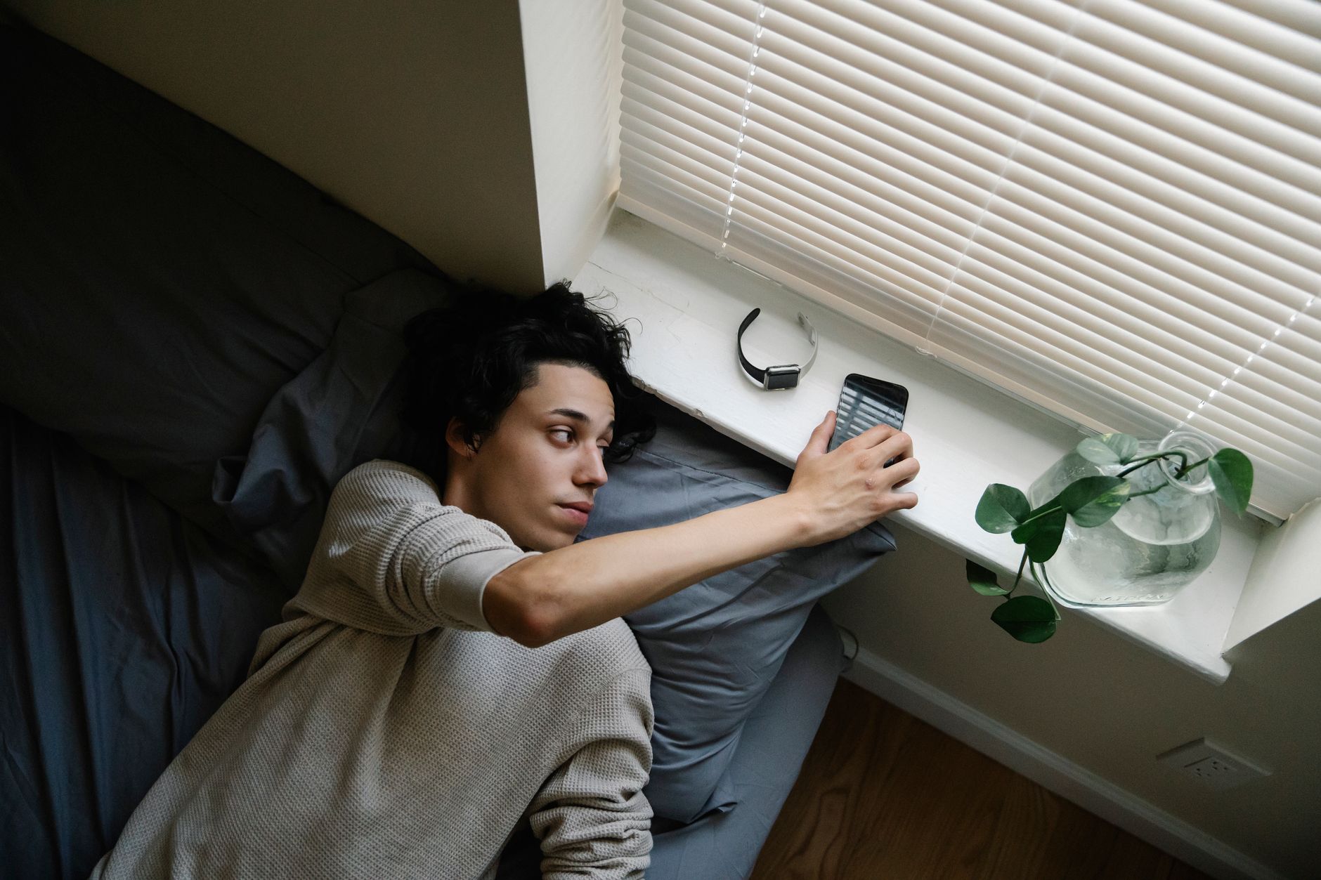 A young man lies on a bed reaching for his phone near a window — the suspended animation of adulthood in a childhood room.