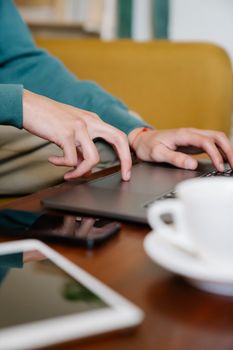 Close-up of hands typing on a laptop in a comfortable indoor space, with coffee and tablet nearby.