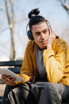 Man sitting outdoors on a bench, using a tablet and wearing headphones, enjoying a peaceful day in the park.