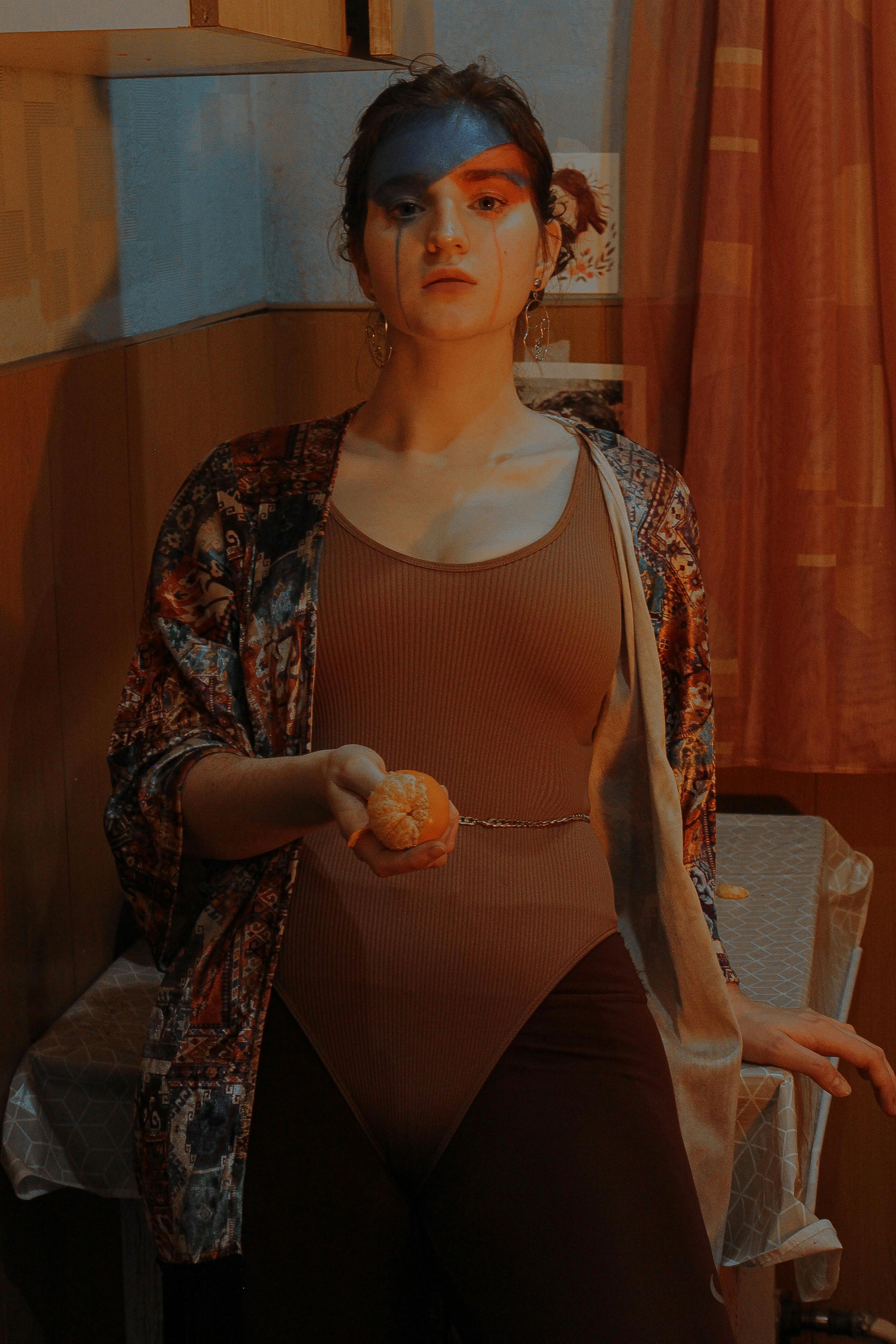 Young female in bodysuit with mascara on cheeks and ripe mandarin looking at camera in room