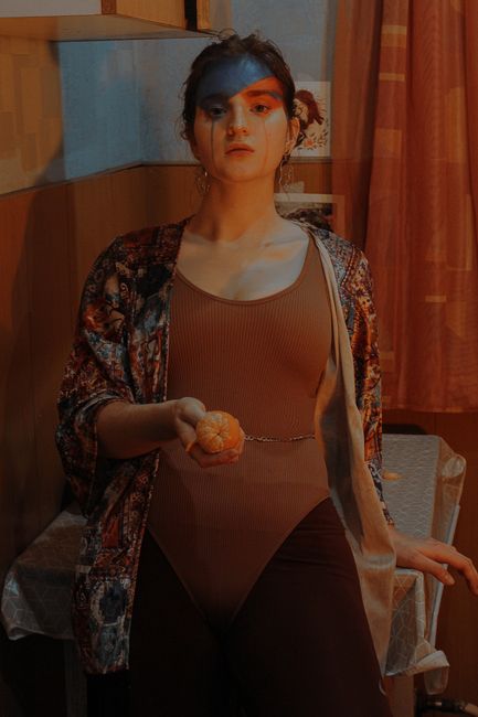 Young female in bodysuit with mascara on cheeks and ripe mandarin looking at camera in room