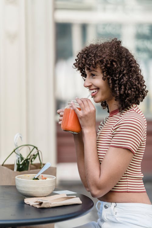 Side view of cheerful ethnic female enjoying tasty refreshing drink while looking forward at cafeteria table with lunch in town