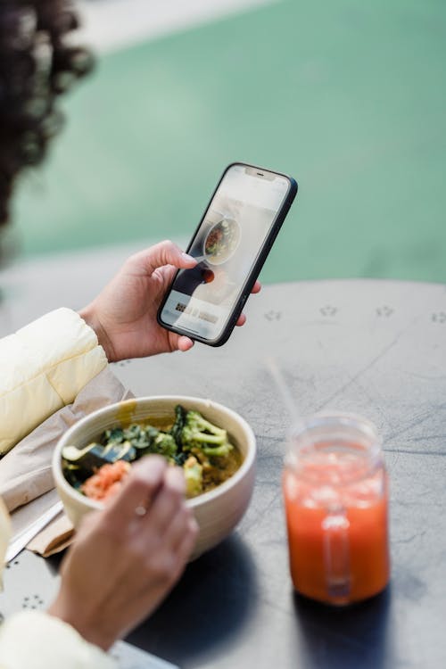 Crop unrecognizable female blogger touching screen on cellphone above street cafe table with healthy food and smoothie