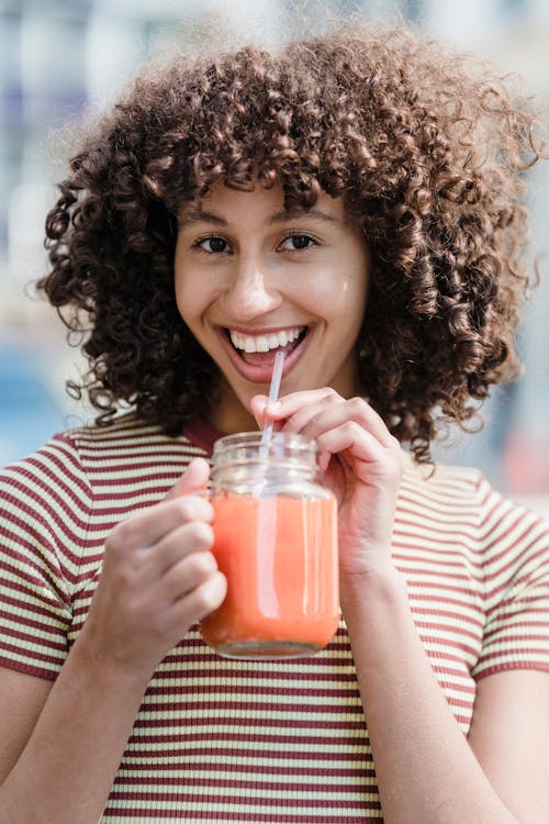 Cheerful ethnic female in striped wear looking at camera while enjoying tasty healthy drink from jar in street cafeteria