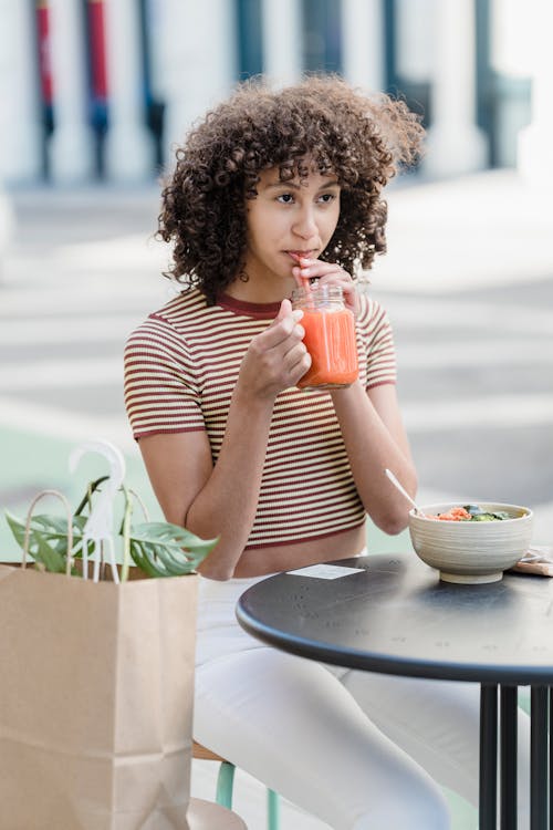 Dreamy young ethnic female enjoying healthy drink while sitting at urban cafeteria table with meal and looking forward