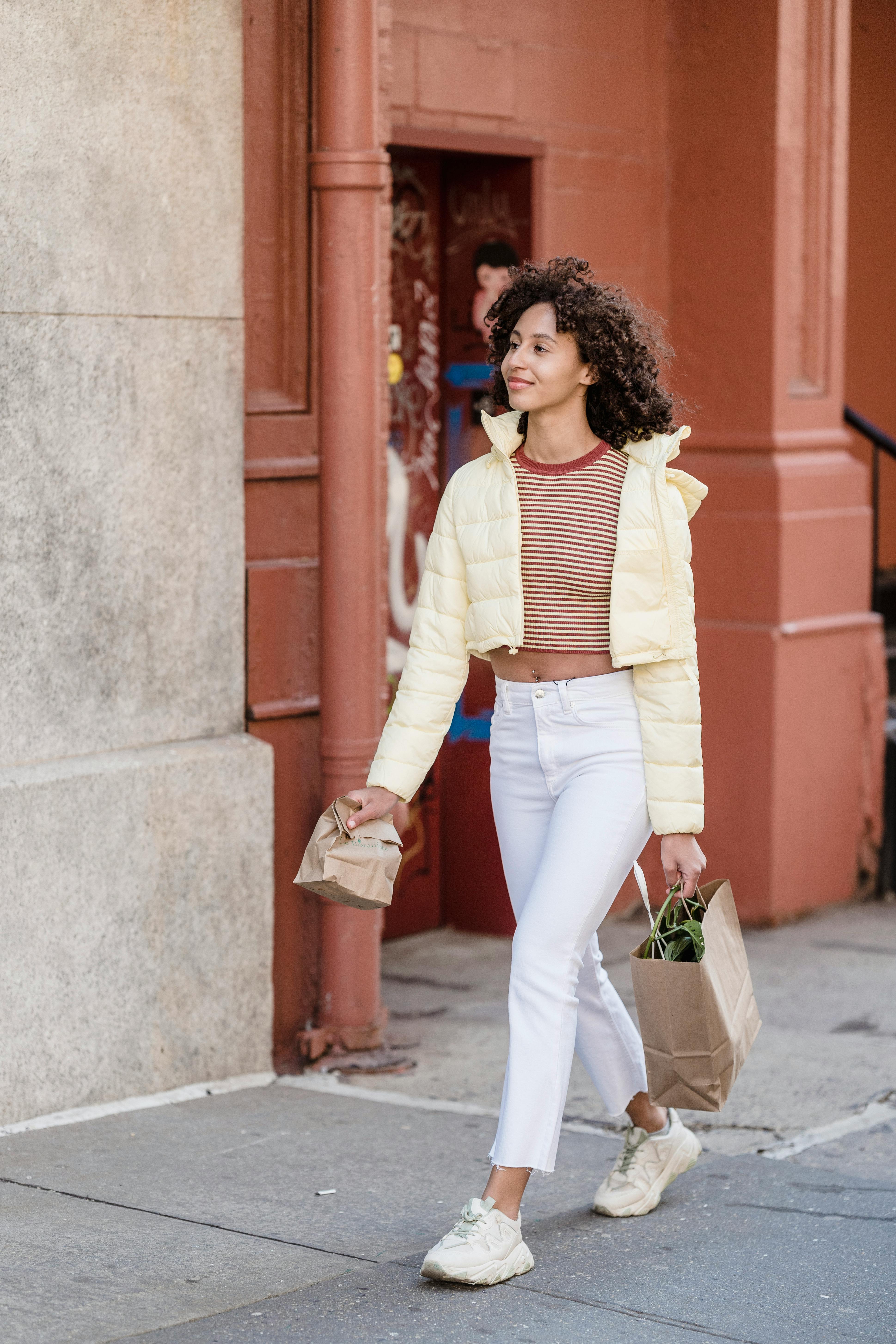 Content young ethnic female buyer with disposable bags strolling on walkway while looking away in town
