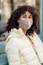 Dreamy young ethnic female in padded jacket and cloth face mask looking away during COVID 19 pandemic in town