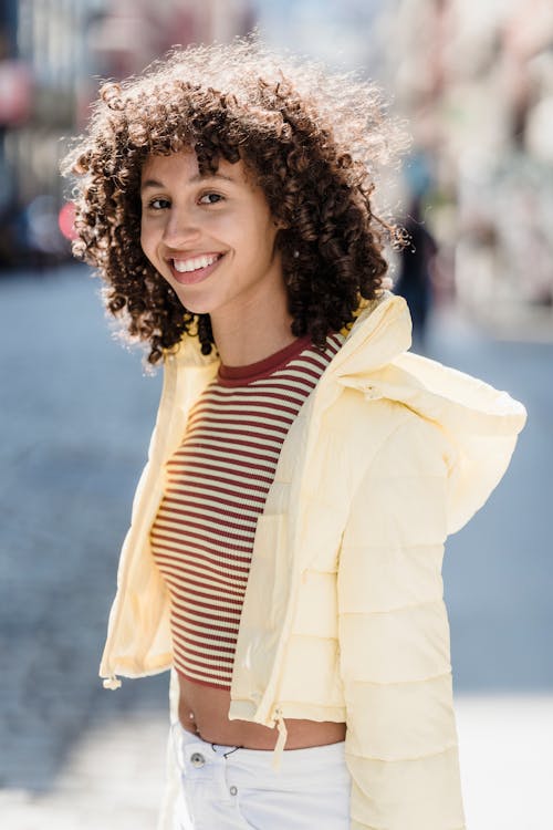 Positive young ethnic female millennial with curly hair in trendy outfit smiling and looking at camera while standing on city street on sunny day