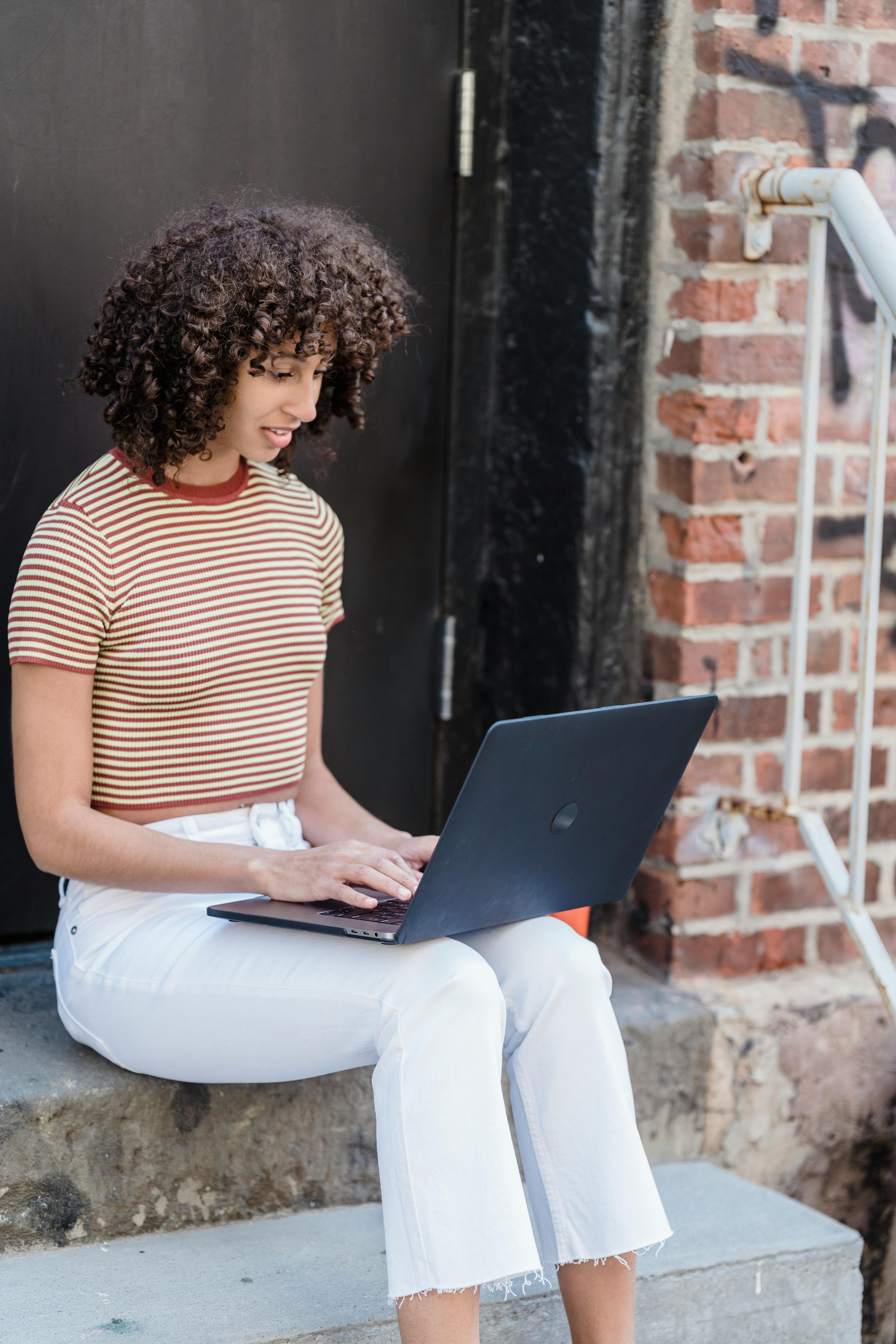 young ethnic woman working online on netbook while sitting on outdoors house stairs