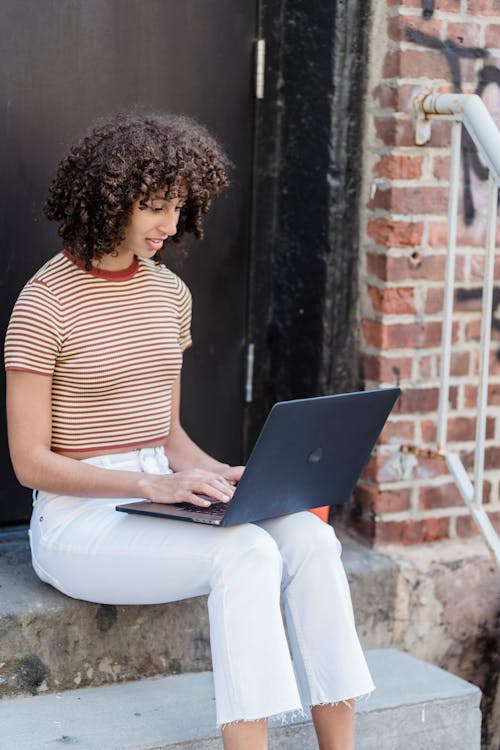Concentrated young stylish ethnic female freelancer with curly hair in casual outfit sitting on stone steps on porch of house and working remotely on laptop