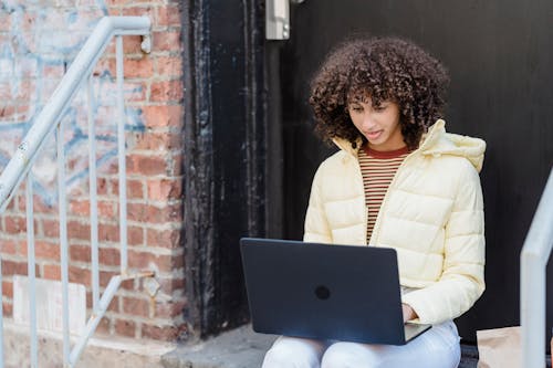 Young ethnic self employed lady with curly hair in trendy outfit sitting on stairs of aged brick building and typing on laptop