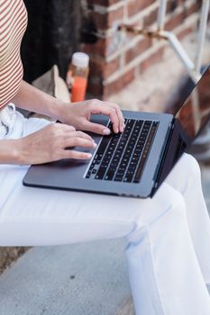 From above of crop anonymous female freelancer in casual clothes typing on netbook while sitting on stairway of aged brick building on street