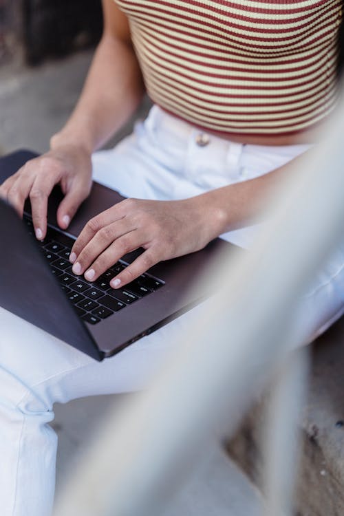 Crop unrecognizable female browsing internet on modern netbook while sitting on street near blurred railing during online work in city