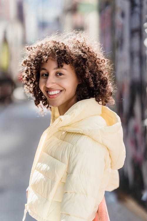 Free Positive ethnic female with curly hair wearing outerwear smiling and looking at camera while standing on blurred street with buildings Stock Photo