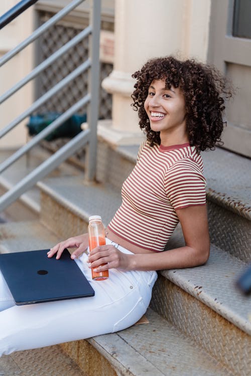 Side view of positive ethnic female in stylish outfit sitting on steps of building with netbook and bottle of juice