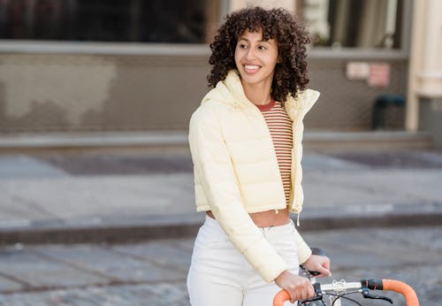 Free Positive ethnic female with curly hair looking away while standing on paved walkway with bicycle on street with building in city Stock Photo