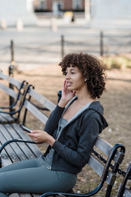 Side view of ethnic female in sportswear listening to music in wireless earbuds while browsing cellphone on bench on blurred street