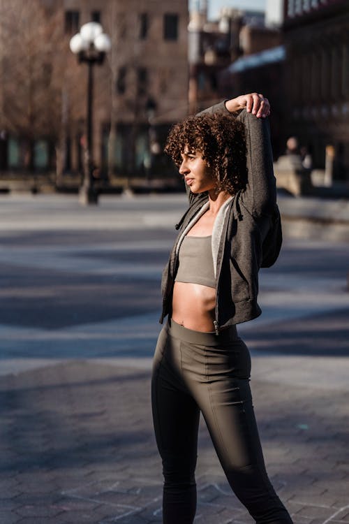 Free Side view of determine ethnic female in activewear doing warm up exercise while standing on paved street with building during work out Stock Photo