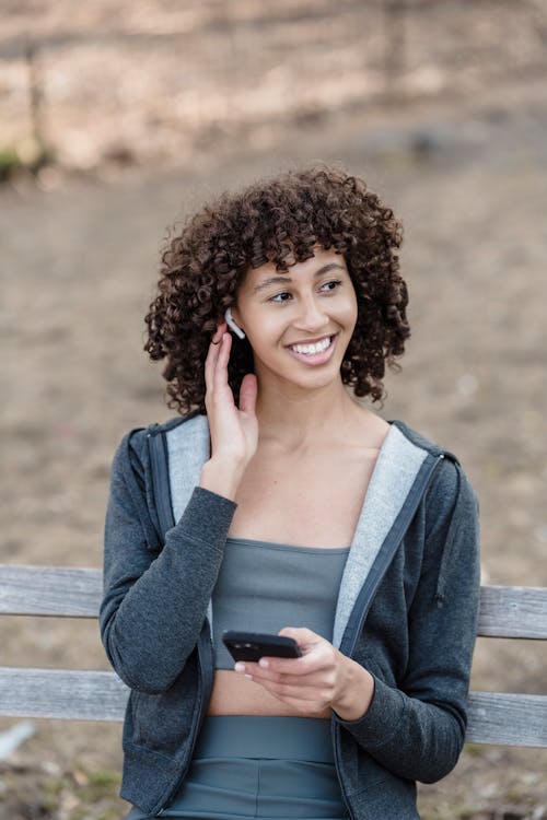 Joyful ethnic female in sportswear looking away and listening to songs in wireless earbuds while browsing smartphone on blurred background