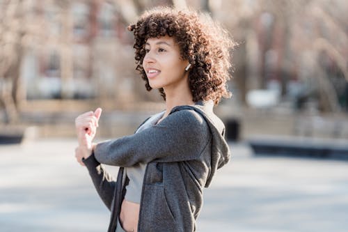 Side view of positive ethnic female doing warm up exercise while listening to music in wireless earbuds on blurred background