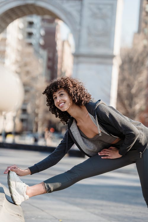 Cheerful ethnic female with curly hair in sportswear and sneakers stretching legs while standing on sunny street with buildings in city