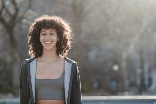 Delighted ethnic sportswoman in activewear in wireless earbuds looking at camera while standing on street against blurred background during training