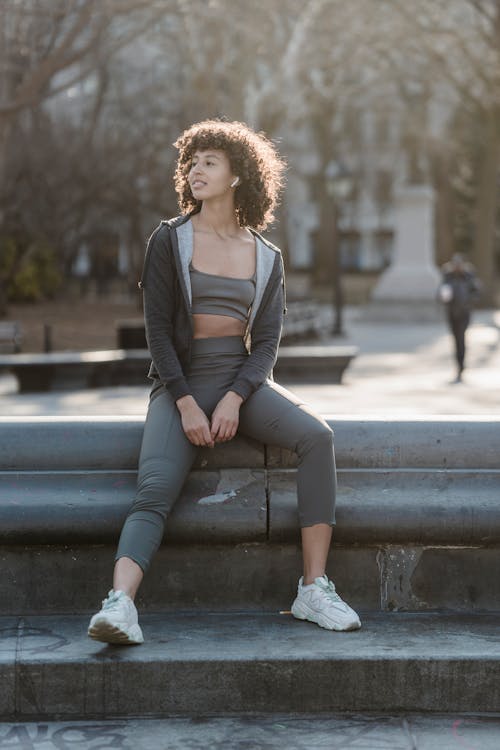 Full body of sporty ethnic female in activewear listening to music in wireless earbuds while sitting on street during training