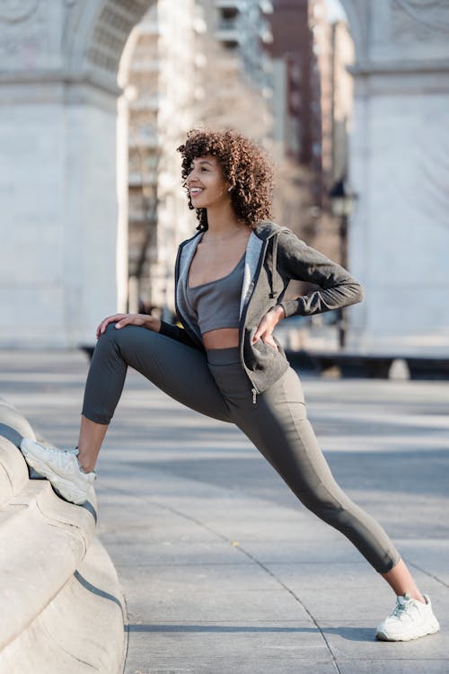 Free Full body side view of happy ethnic female in activewear warming up while standing near border on street with historic arch Stock Photo