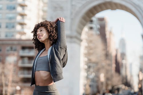 Free Curly haired toned ethnic female in activewear doing warming up exercise while training on city street Stock Photo