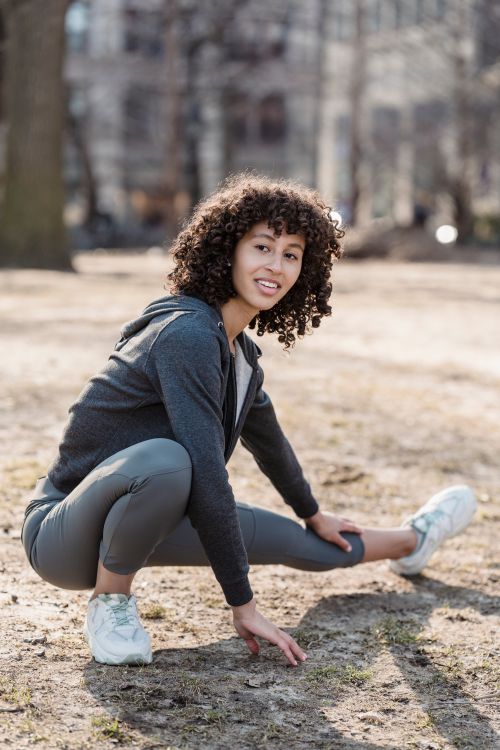 Free Full body of positive ethnic female runner with curly hair stretching legs while warming up during fitness training in park Stock Photo