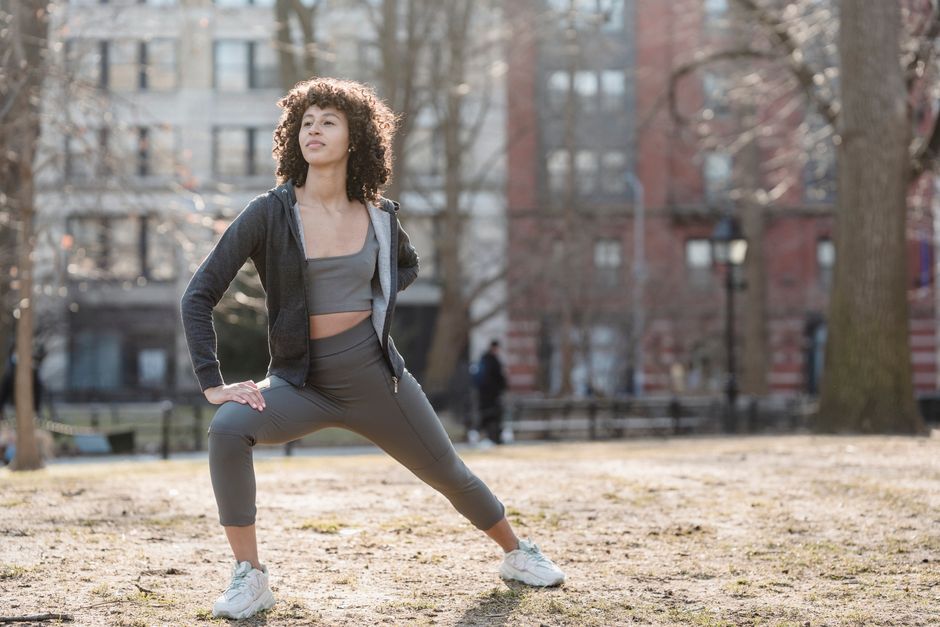 Full body of black female in sportswear stretching legs during fitness workout in park on blurred background and looking away