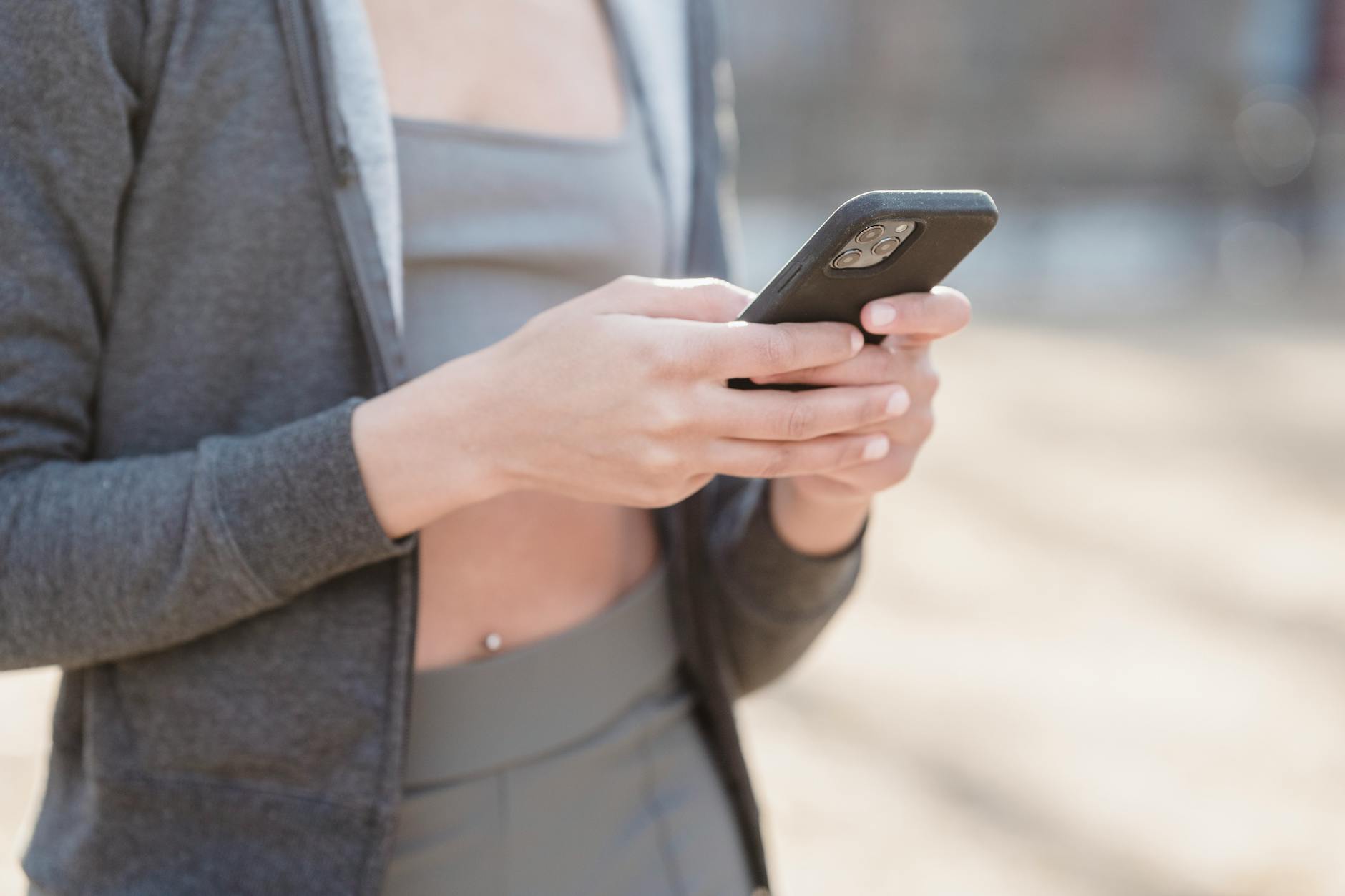 Crop anonymous female in gray outfit using mobile phone on street on blurred background