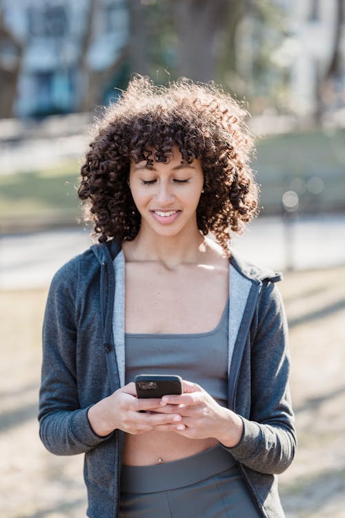 African American female with curly hair smiling on street while browsing mobile phone in daylight