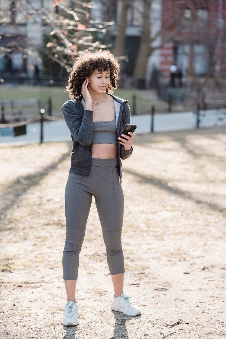 Black Woman Touching Ear And Looking At Smartphone In Park