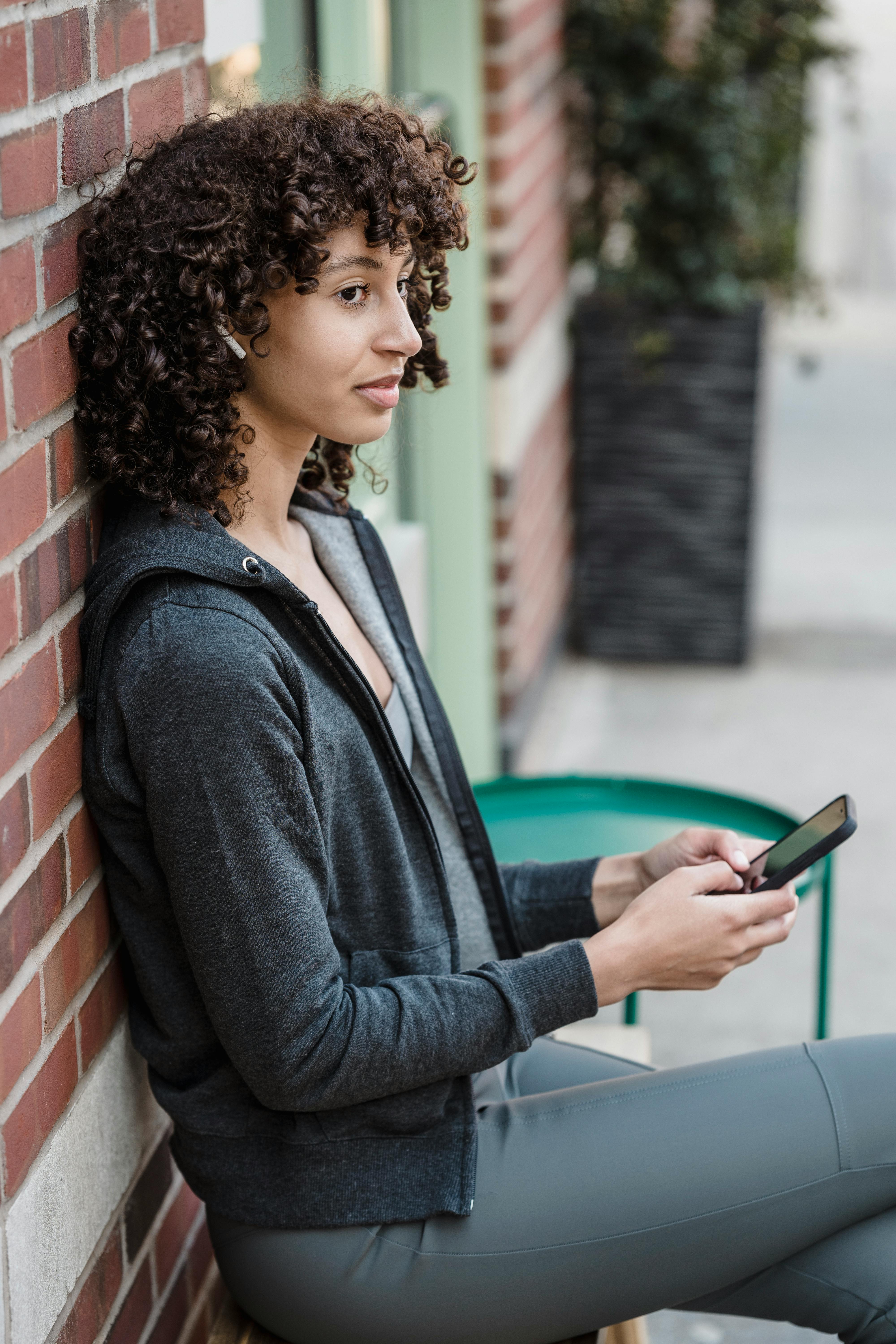 young ethnic woman in tws earphones using smartphone on bench in city